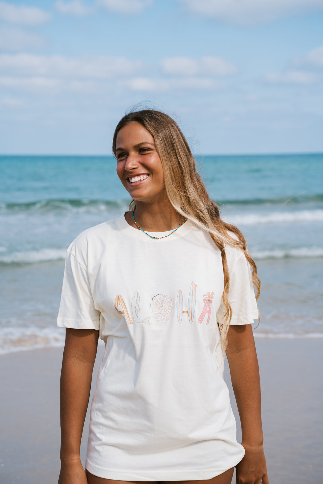אשה בחוף הים לובשת טי שירט לבנה עם הכיתוב "Aloha" – Lika Swimwear