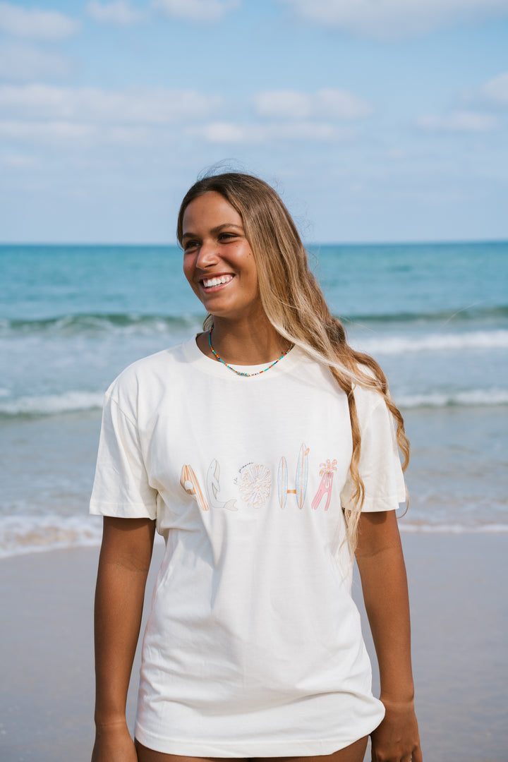 אשה בחוף הים לובשת טי שירט לבנה עם הכיתוב "Aloha" – Lika Swimwear