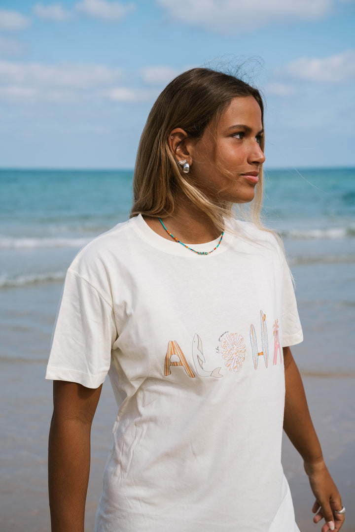אשה בחוף הים לובשת טי שירט לבנה עם הכיתוב "Aloha" – Lika Swimwear
