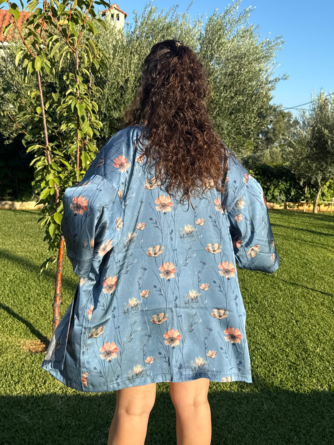 בחורה לובשת שמלת פרחים כחולה של Lika Swimwear בעמידה באזור דשא עם עצים, מראה קז’ואל ורענן.