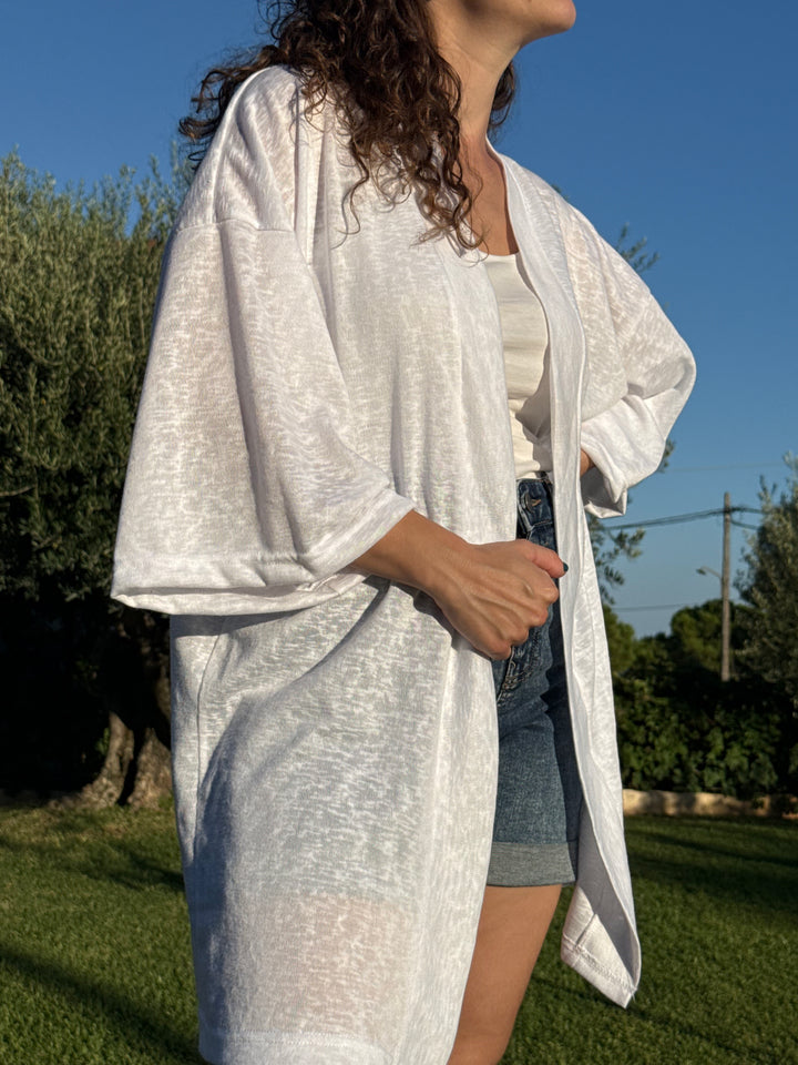אשה לובשת קימונו לבן בסגנון Lika Swimwear בחוץ, עם רקע ירוק וטבעי, מראה קז’ואל אלגנטי ונוח.