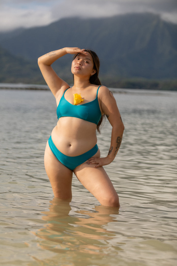 אישה עם גוף מלא בביקיני טורקיז מבית Lika Swimwear עומדת במים, בגדי ים לנשים, נוף הררי של הוואי ברקע
