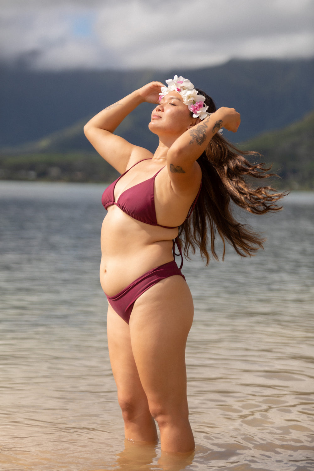 אישה עם גוף מלא לובשת בגד ים אדום של LIKA Swimwear עם סרט פרחים בשיער, עומדת ליד מים עם הרים ברקע.