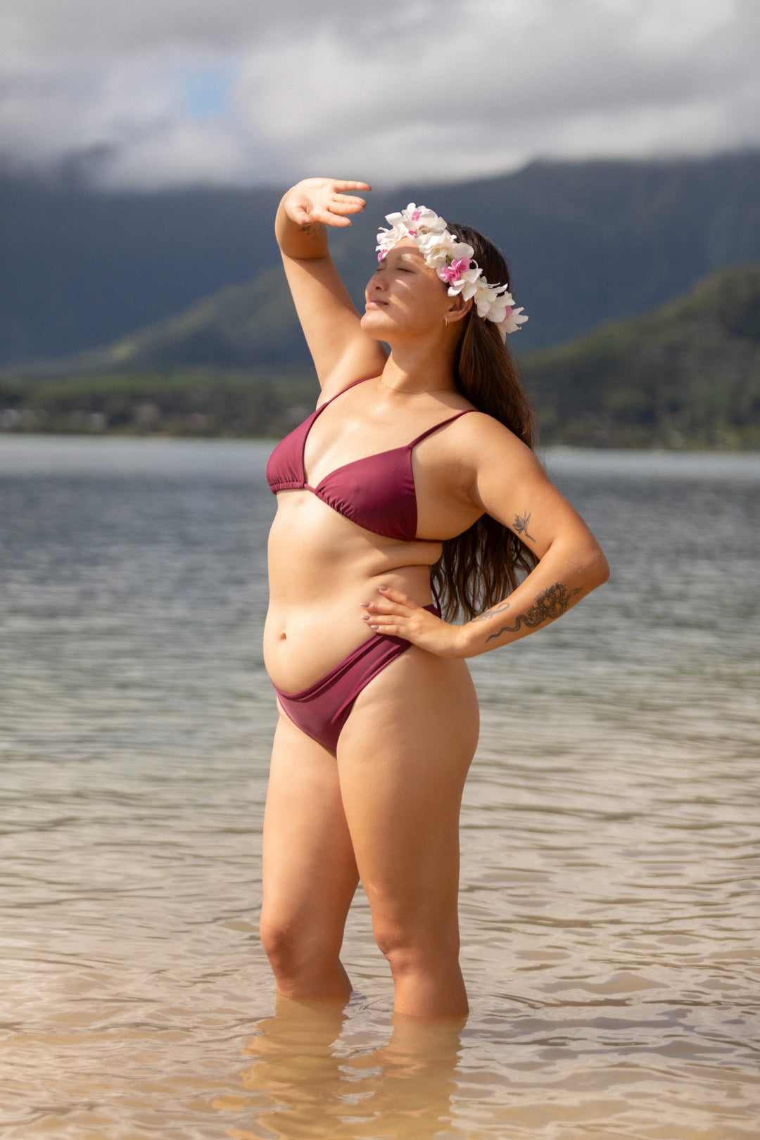 אישה עם גוף מלא לובשת בגד ים אדום של LIKA Swimwear עם סרט פרחים בשיער, עומדת ליד מים עם הרים ברקע.