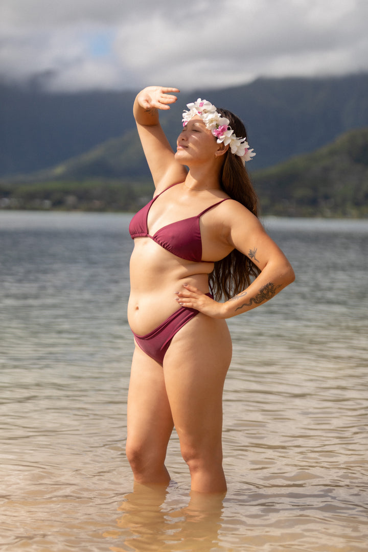אישה עם גוף מלא לובשת בגד ים אדום של LIKA Swimwear עם סרט פרחים בשיער, עומדת ליד מים עם הרים ברקע.